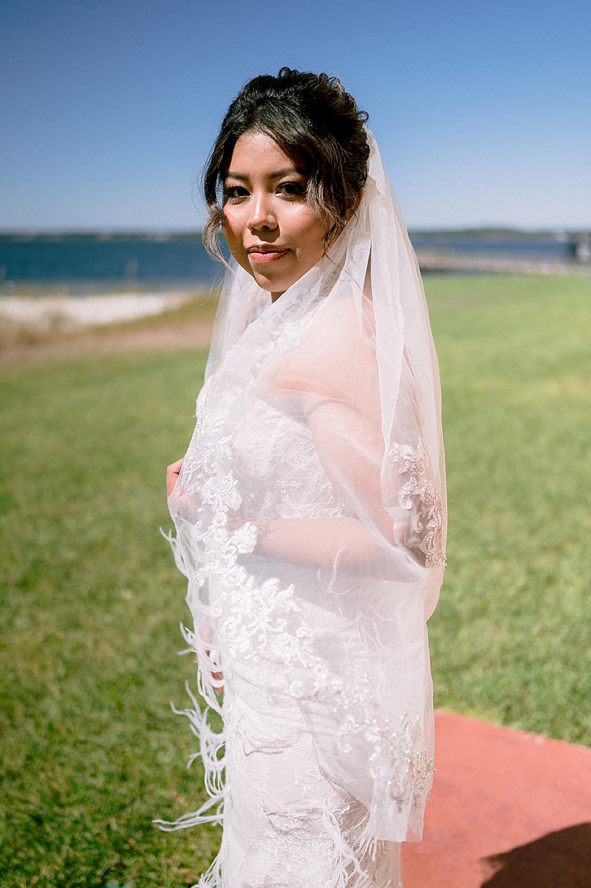 bride holds veil over arm on beach before ceremony by Emily burns photo